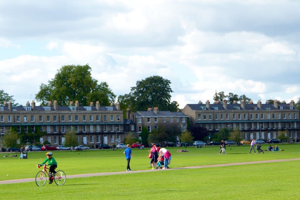 A park with a green lawn, a person riding a bicycle, and a row of houses in the background.