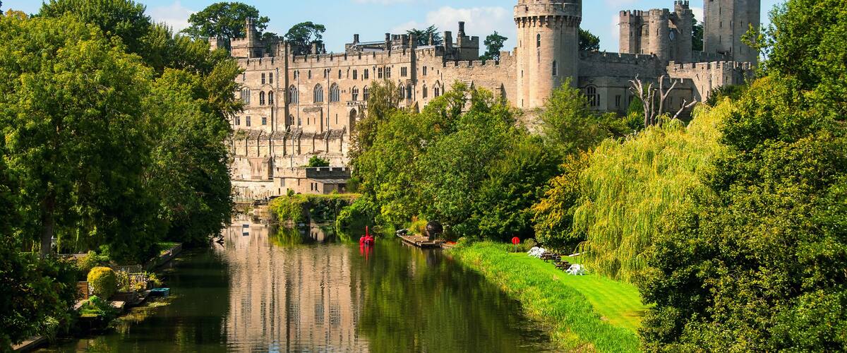 A castle with a river in the foreground and lush greenery.