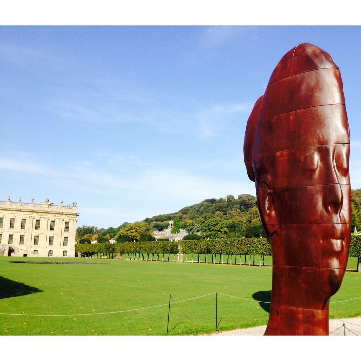 A large bronze sculpture of a man's head in a park with a historic building in the background.