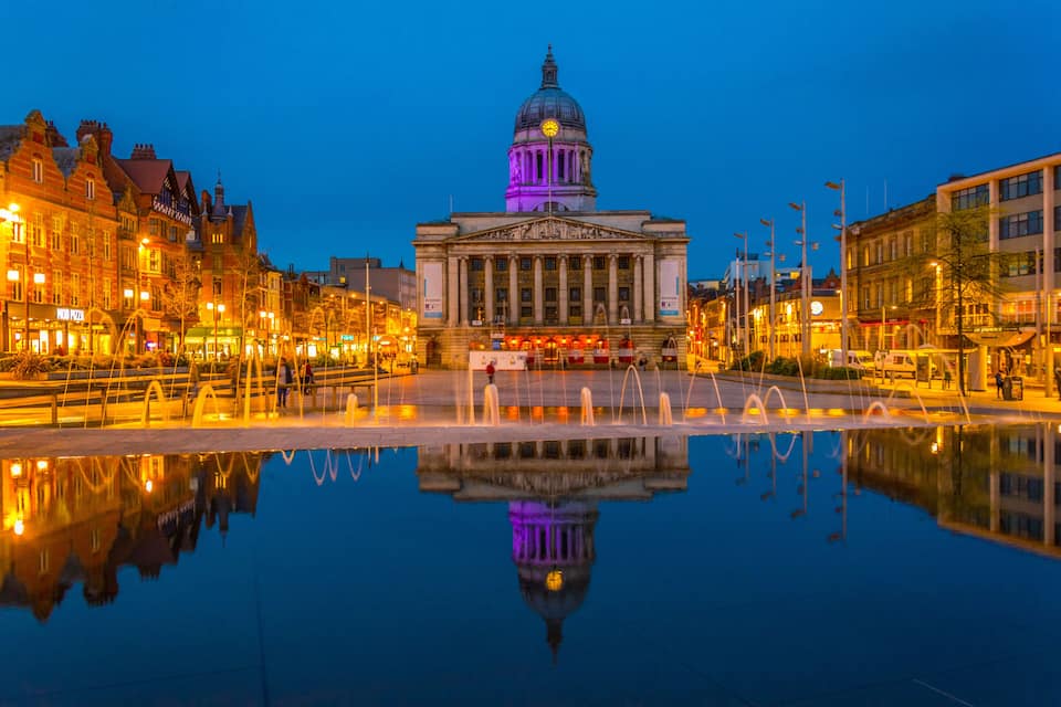 A historic building with a dome and columns, reflecting in a calm water body surrounded by illuminated buildings.