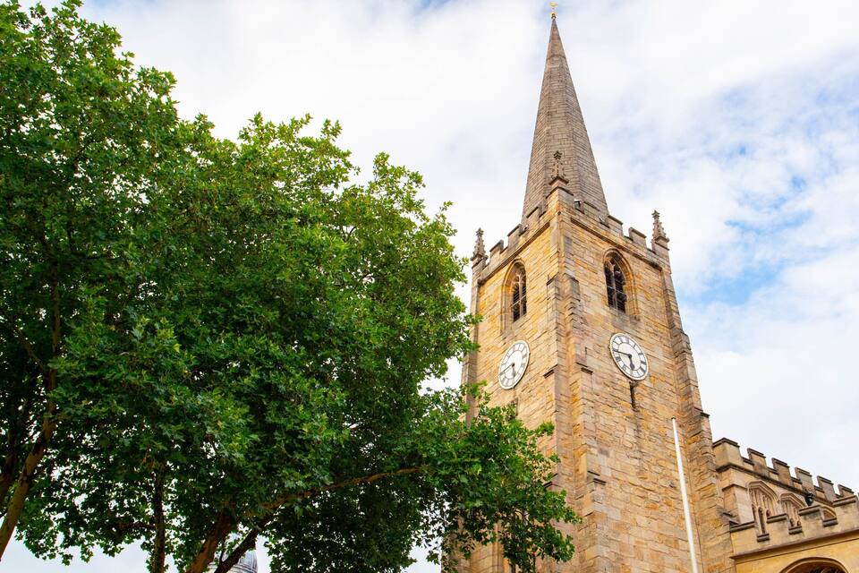 Een hoge stenen toren met een puntige spits en twee klokken, omgeven door weelderige groene bomen.