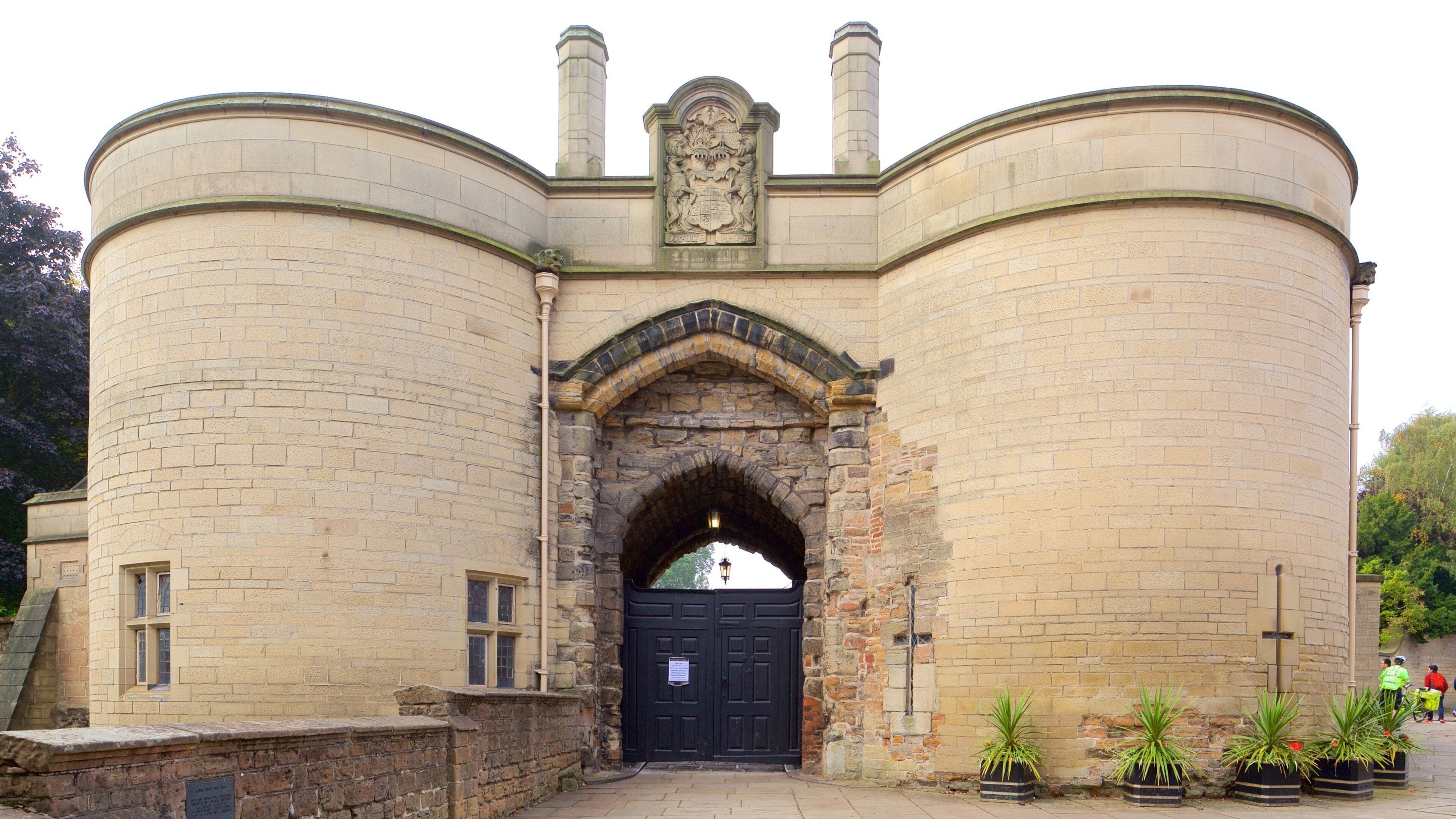 A historic stone building with a black arched entrance, a coat of arms above the door, and potted plants in front.