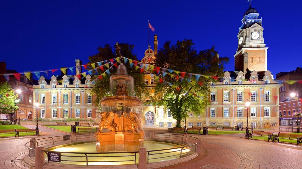 A historic building with a clock tower, a fountain, and a well-lit square with benches.