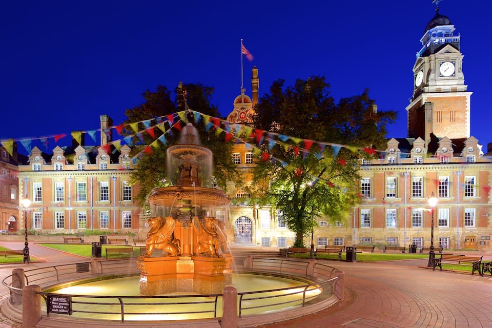 A historic building with a clock tower, a fountain, and a well-lit square with benches.