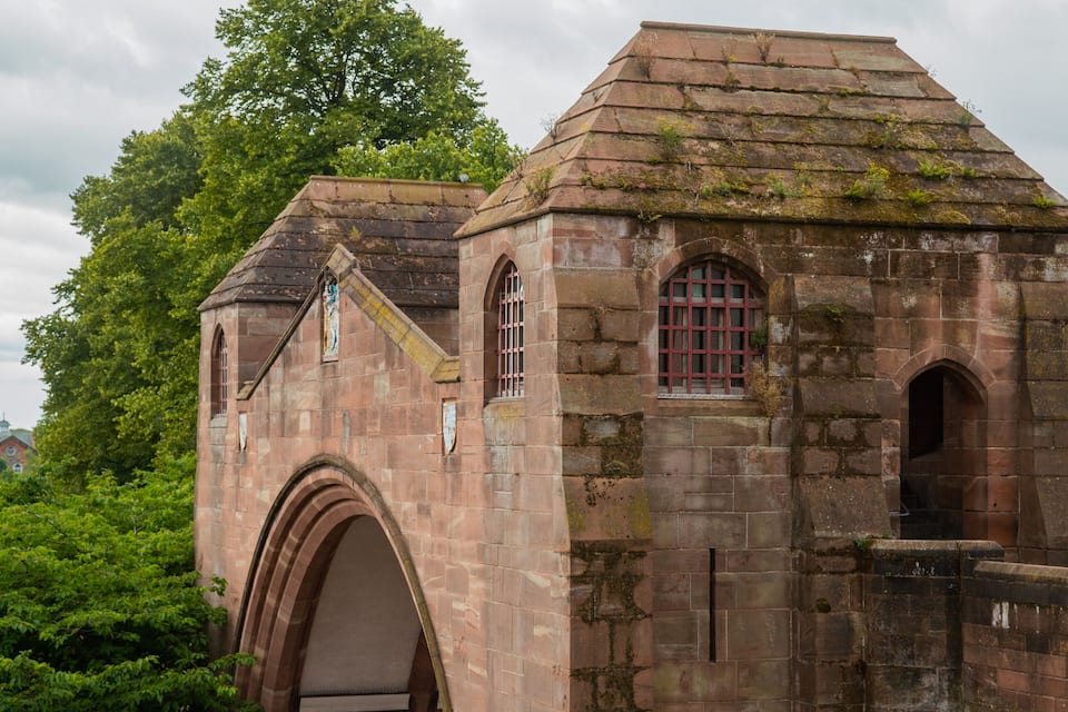 A historic stone building with arched windows and a tiled roof, surrounded by lush greenery.