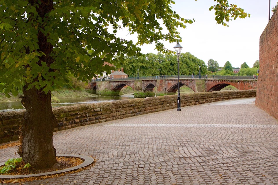 A tree-lined cobblestone path beside a river, with a brick bridge in the background.