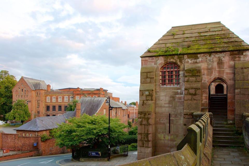 A historic stone tower with a pointed roof, a brick building with arched windows, and a street with a pedestrian crossing.