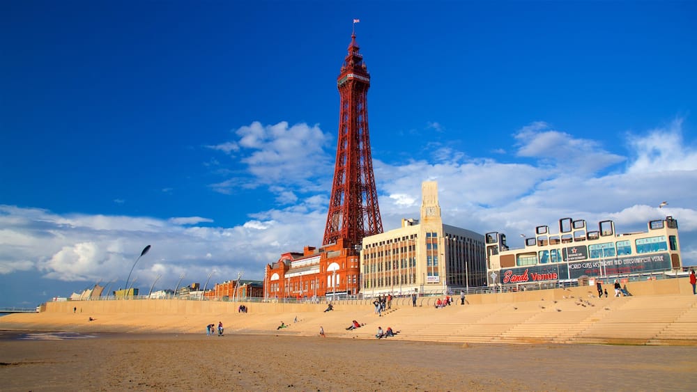 A seaside promenade with a prominent red tower, a large building with a clock tower, and a double-decker bus.