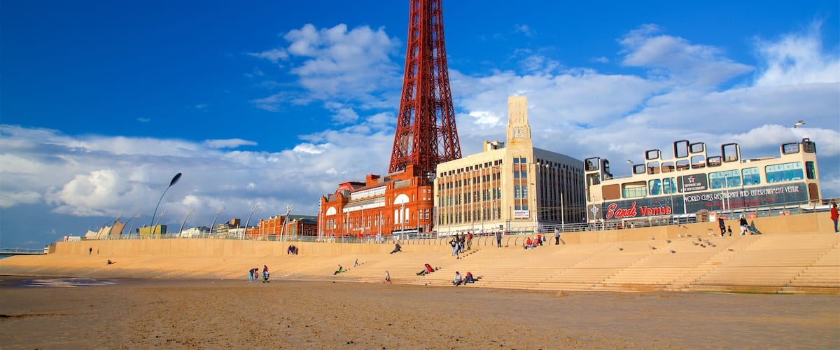 A seaside promenade with a prominent red tower, a large building with a clock tower, and a double-decker bus.