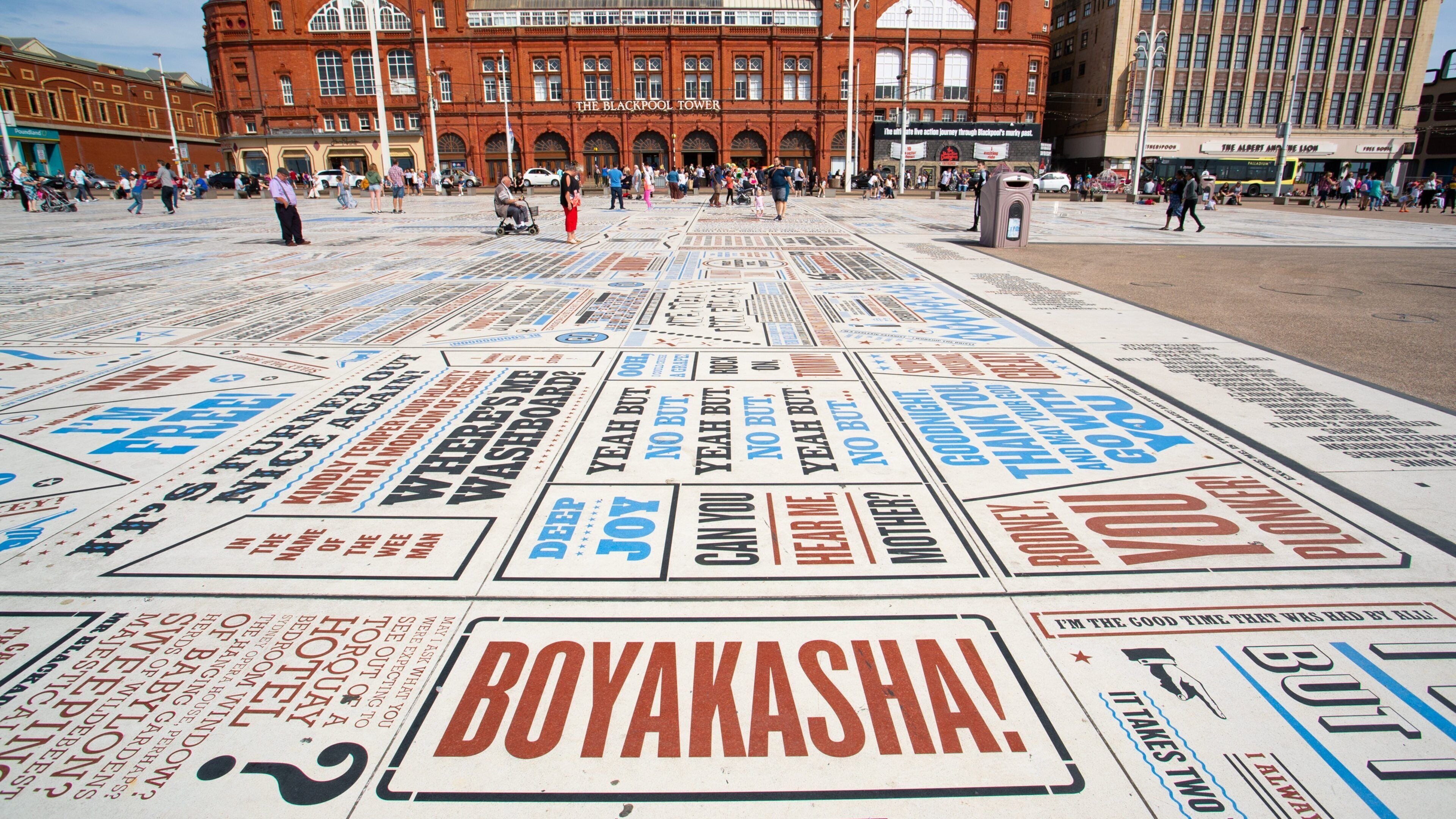 Una plaza pública con una gran instalación colorida que presenta varias frases y nombres, incluyendo 'BOYAKSHA!' en primer plano.