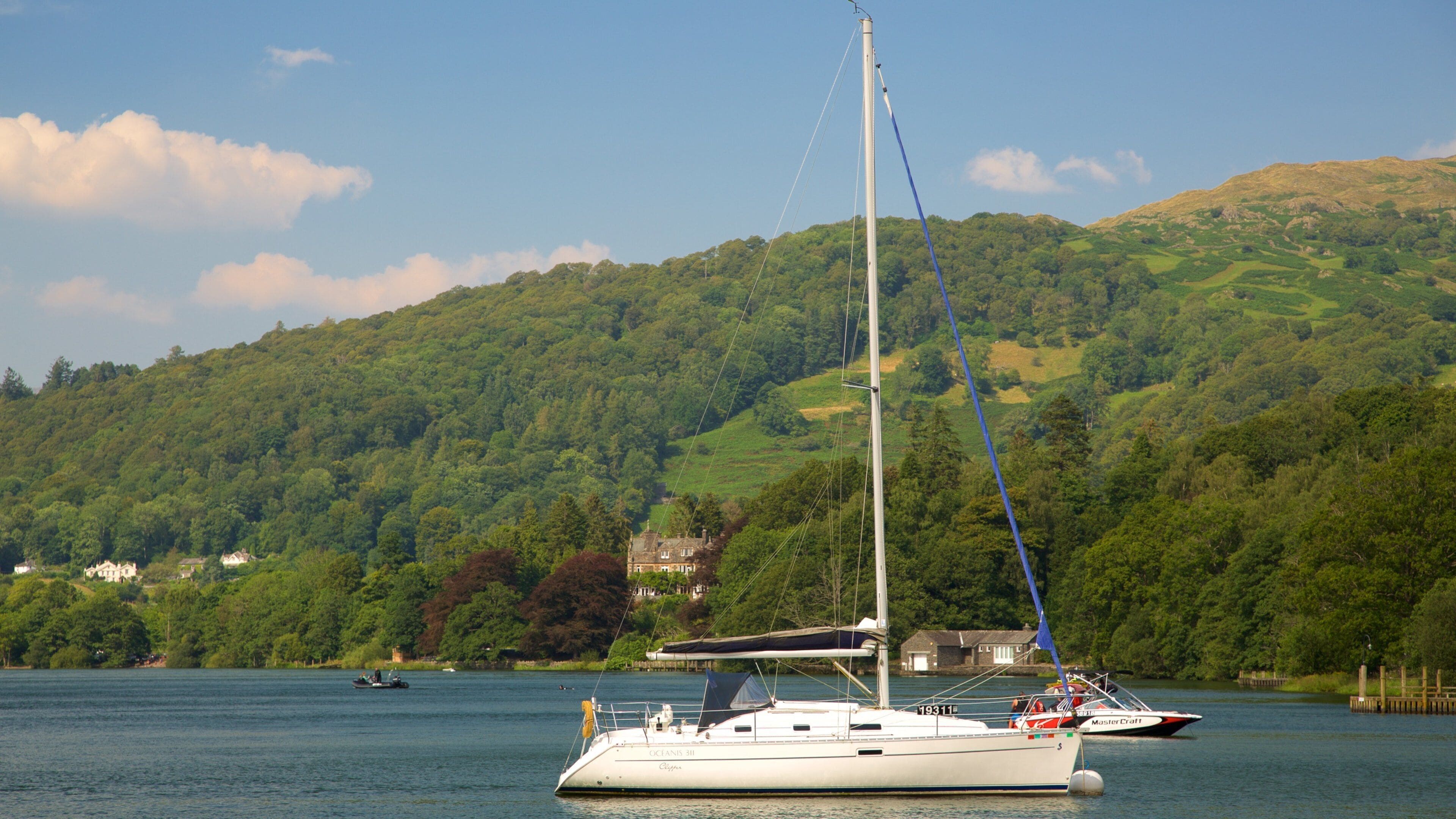 A sailboat named 'Oceans 31' on a calm lake with a backdrop of lush green hills and a few houses.