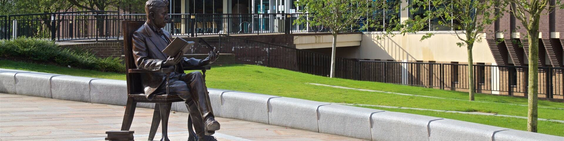 A statue of a man reading in a park with a building labeled "The Pottery Museum & Art Gallery" in the background.