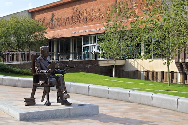 Une statue représentant un homme lisant dans un parc, avec un bâtiment portant l’inscription « Le musée de la céramique et la galerie d’art » en arrière-plan.