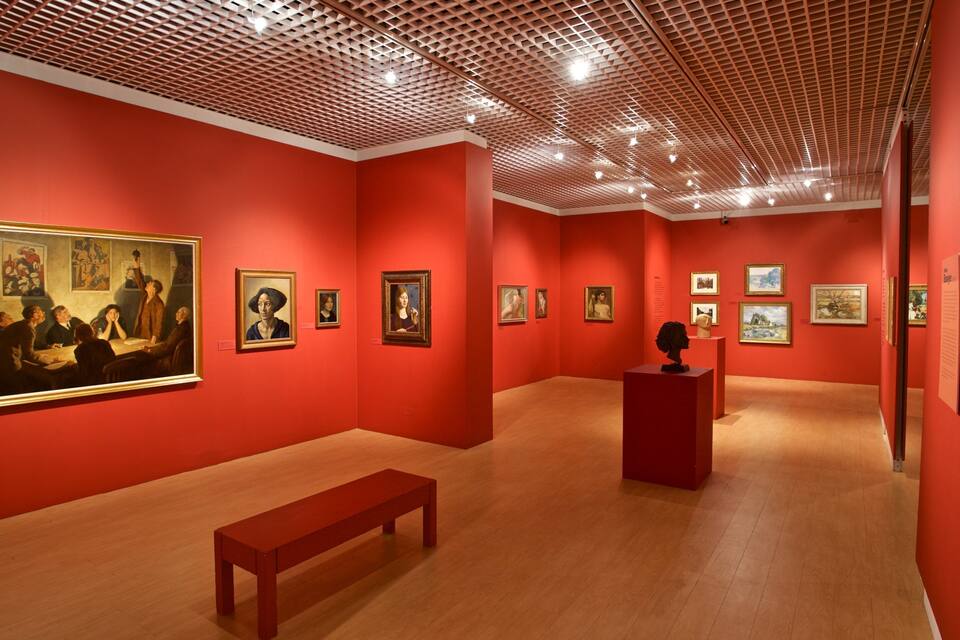 An art gallery with red walls and wooden floors, featuring various paintings and sculptures.