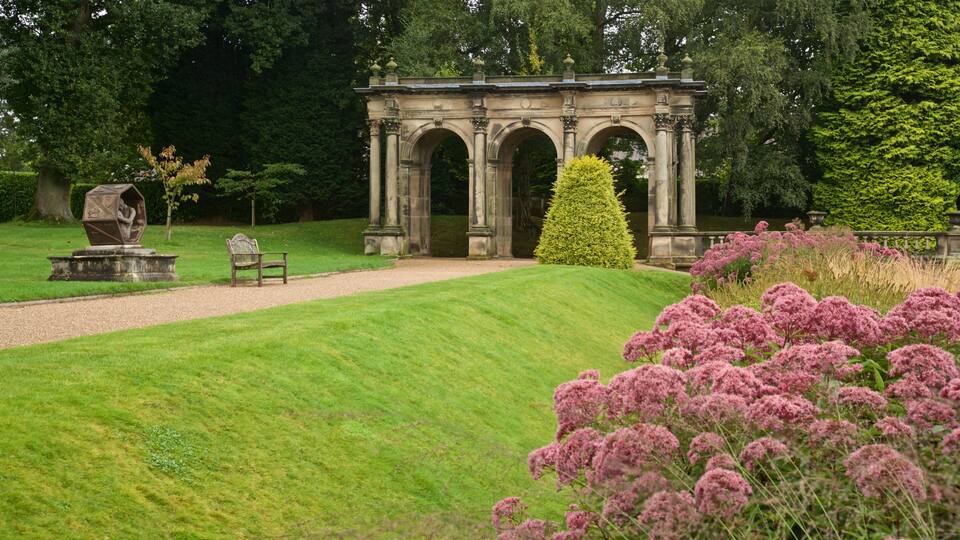 A classical stone structure with arches, a neatly trimmed lawn, and pink flowering plants.