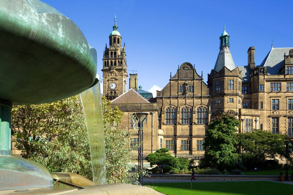 A historic building with a clock tower, a fountain, and lush greenery.