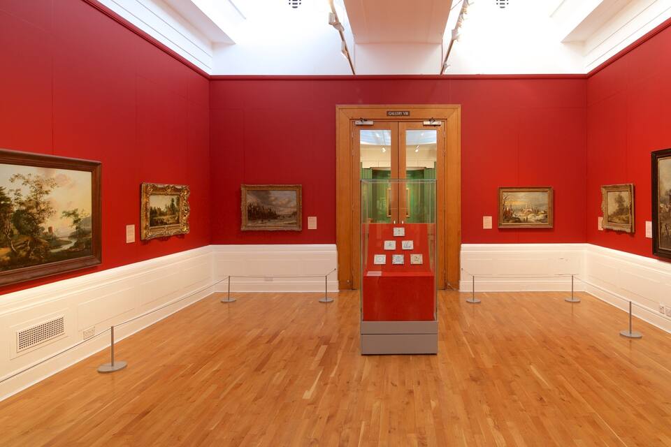 Uma galeria de arte com paredes vermelhas e chão de madeira, exibindo diversas pinturas emolduradas.