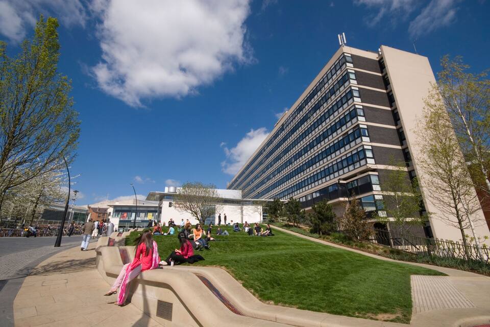 A modern building with a glass facade, a grassy area with people sitting, and a clear blue sky.