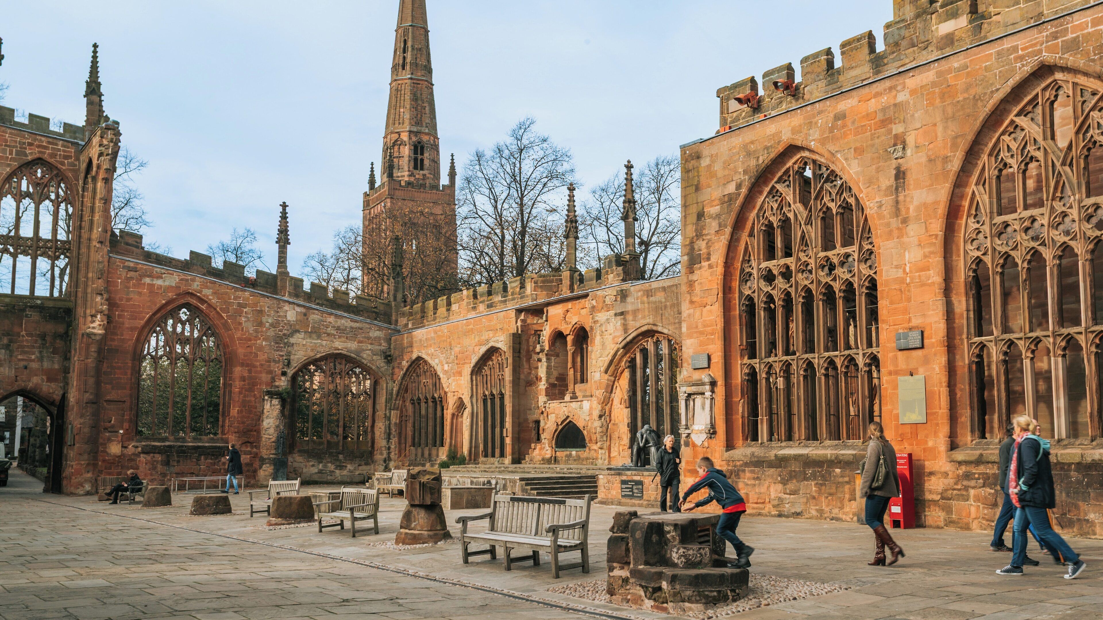 Coventry Cathedral stands in historical ruins, surrounded by visitors exploring its majestic architecture in Coventry City Centre, England on a calm day