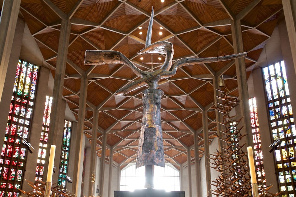 A large metal sculpture in a cathedral with a high, geometric wooden ceiling and colorful stained glass windows.