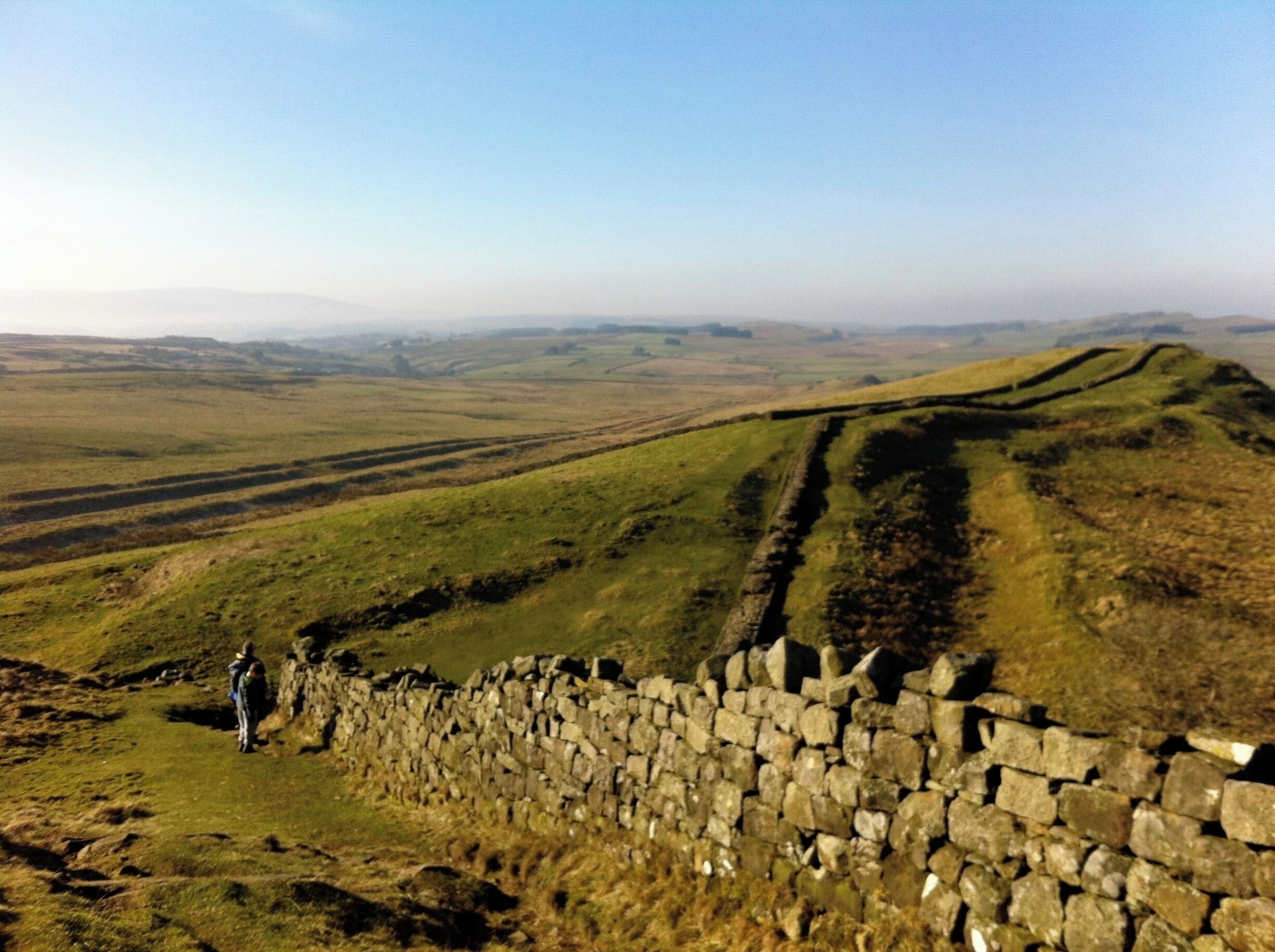 A stone wall with a person standing beside it, overlooking rolling hills and a distant horizon.