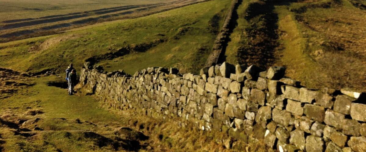 A stone wall with a person standing beside it, overlooking rolling hills and a distant horizon.