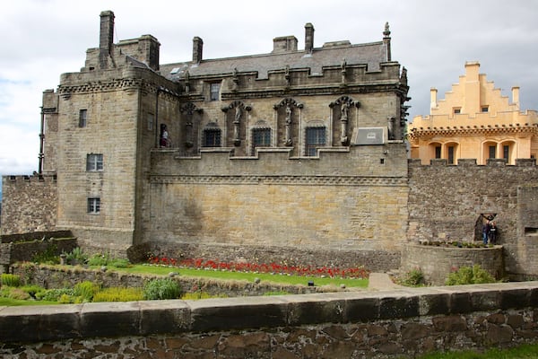 Un château historique doté d’un grand mur de pierre, d’un bâtiment plus petit attenant et d’un jardin bien entretenu agrémenté de fleurs rouges.