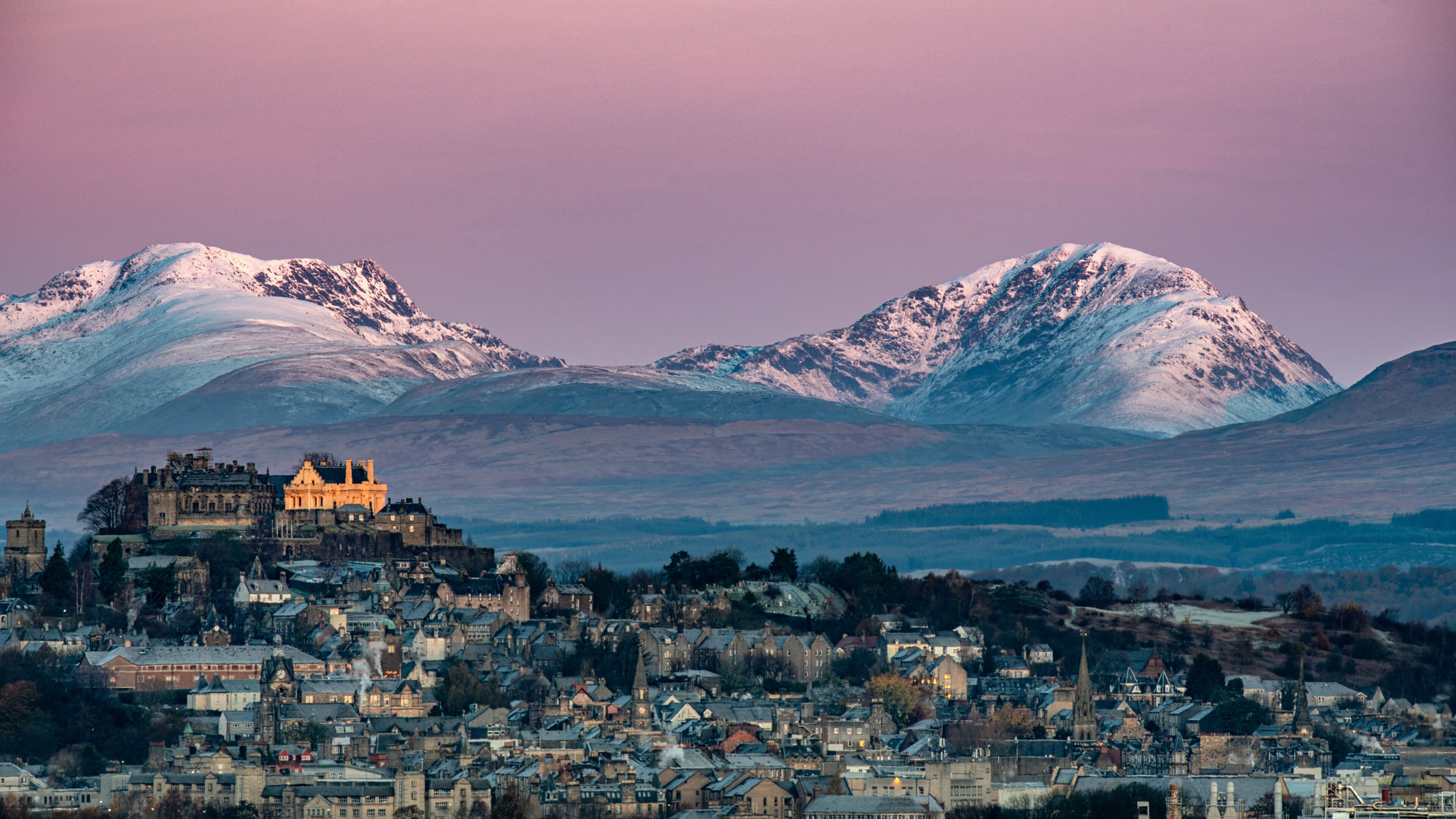 A historic castle overlooking a town with snow-capped mountains in the background.