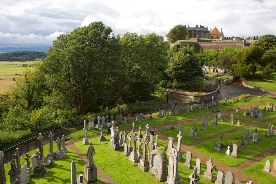 A cemetery with numerous gravestones, surrounded by lush greenery and a castle in the background.