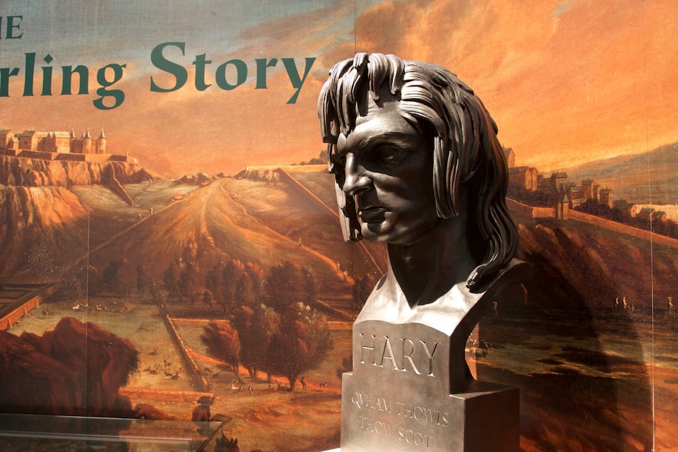 A sculpture of a person with a bust and a bust base labeled "HARRY QVM THOWIS THOWIS SCOT" against a backdrop of a castle on a hill and a landscape with trees and a river.
