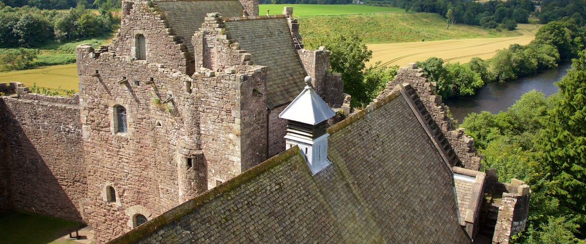 A historic stone castle with battlements, a river, and rolling green hills in the background.