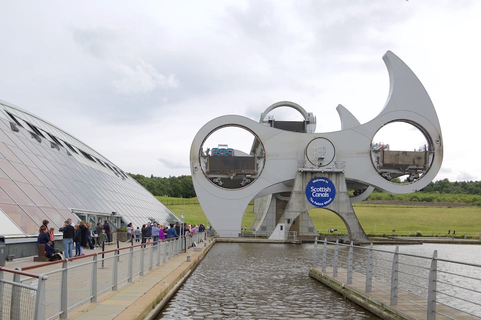 A modern architectural structure with a circular design, a sign reading "Welcome to Scottish Canals," and a body of water.