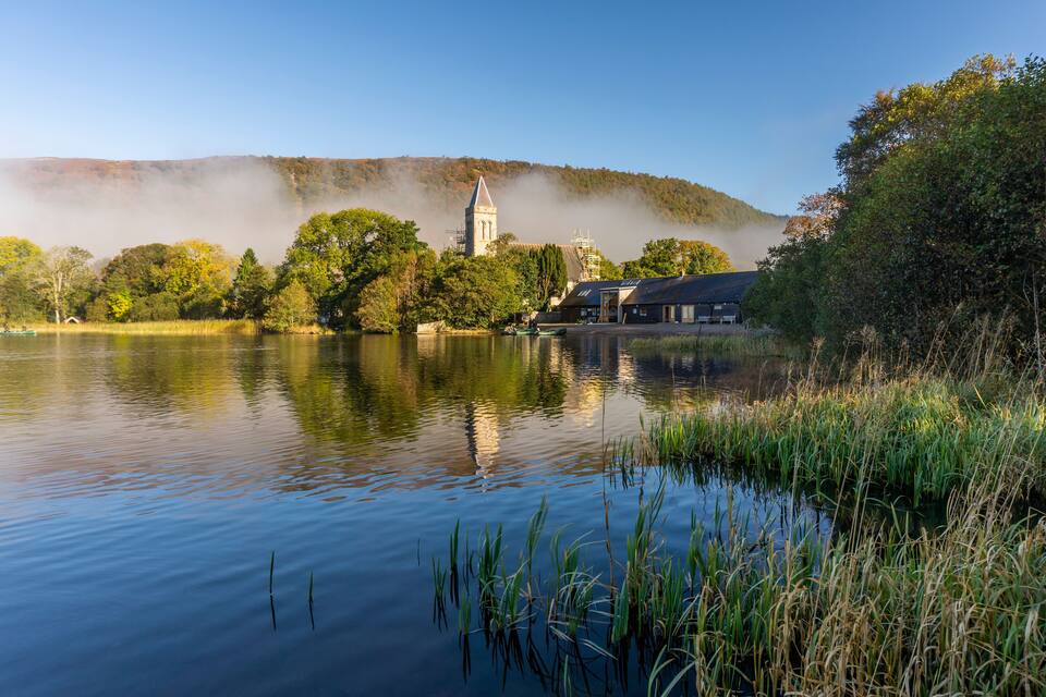 A calm lake with a church and buildings on the shore, surrounded by trees and hills.