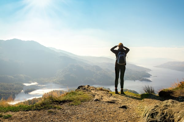 Ein Wanderer auf einem felsigen Vorsprung mit Blick auf ein Tal, einen See und Berge im Hintergrund.