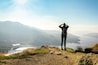 A hiker on a rocky outcrop overlooking a valley with a lake and mountains in the background.