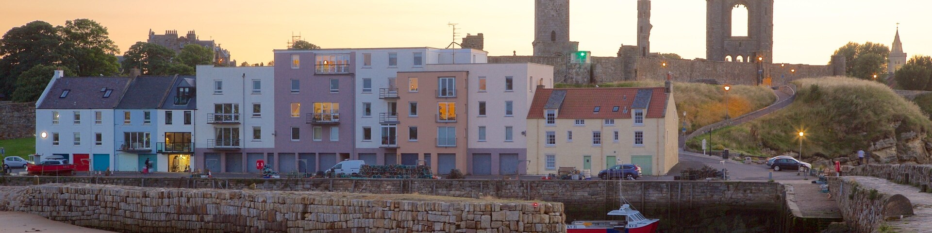 Un pueblo costero con edificios de colores, un muro de piedra, un pequeño bote y una torre histórica al fondo.