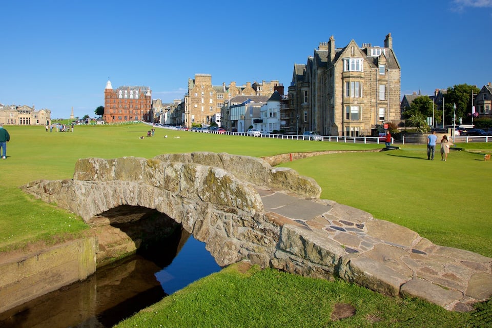 Un puente de piedra sobre un canal, un campo cubierto de césped y edificios históricos al fondo.