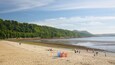 A sandy beach with a forested hillside, a calm sea, and a few people and beach equipment.