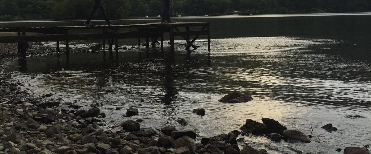 A rocky shoreline with a wooden pier extending into a calm lake, surrounded by green hills.