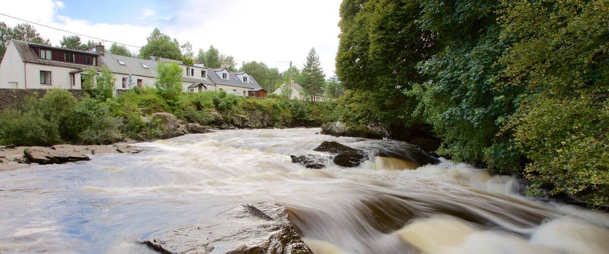 A fast-flowing river with rocks, surrounded by greenery and residential buildings.