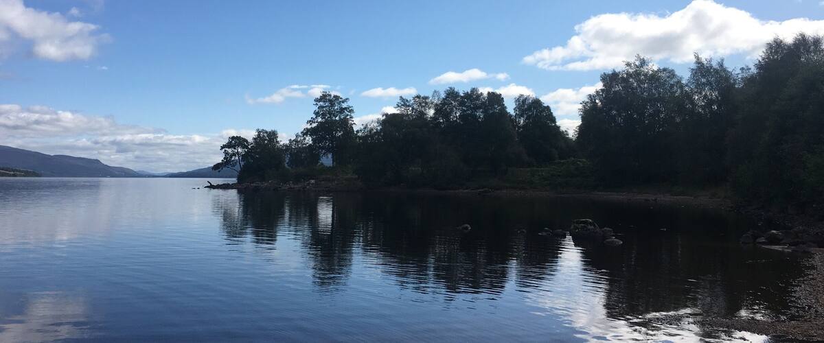 A calm lake with a clear sky, surrounded by trees and distant mountains.
