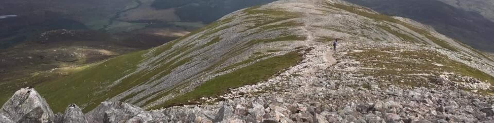 A rugged mountain ridge with a trail, a valley with a lake, and distant mountains under a cloudy sky.