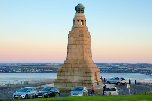 A tall stone monument with a green top, surrounded by parked cars and a waterfront in the background.