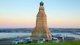 A tall stone monument with a green top, surrounded by parked cars and a waterfront in the background.