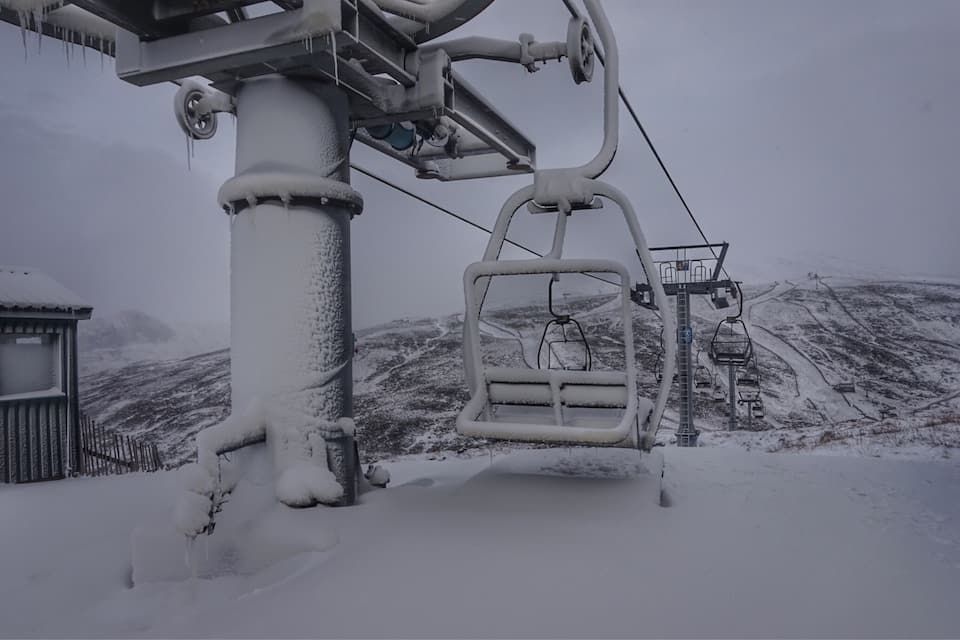 A snow-covered ski lift with a chair in the foreground and a mountain in the background.