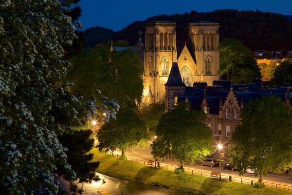 Une église historique à l’architecture gothique, entourée d’arbres et bordée d’une rue bien éclairée au crépuscule.