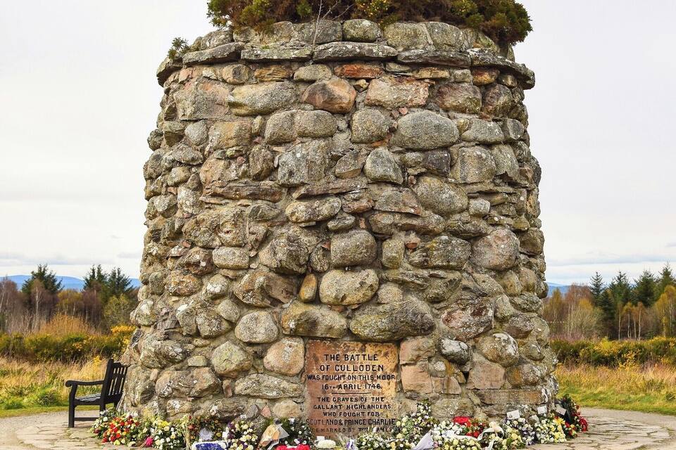 Un monumento in pietra con una targa commemorativa della battaglia di Culloden, circondato da fiori e una panchina.
