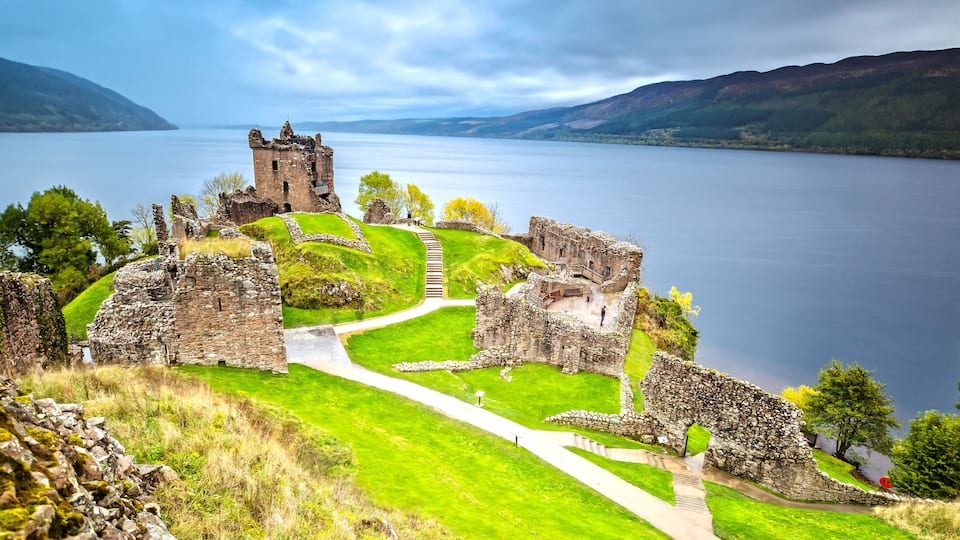 The ruins of a castle overlooking a calm lake with surrounding hills.