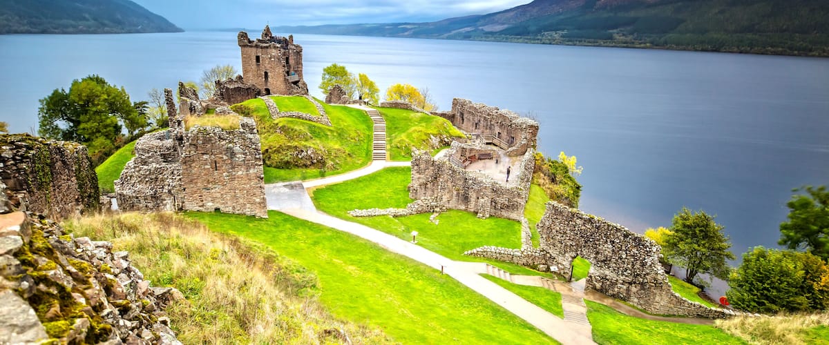 The ruins of a castle overlooking a calm lake with surrounding hills.