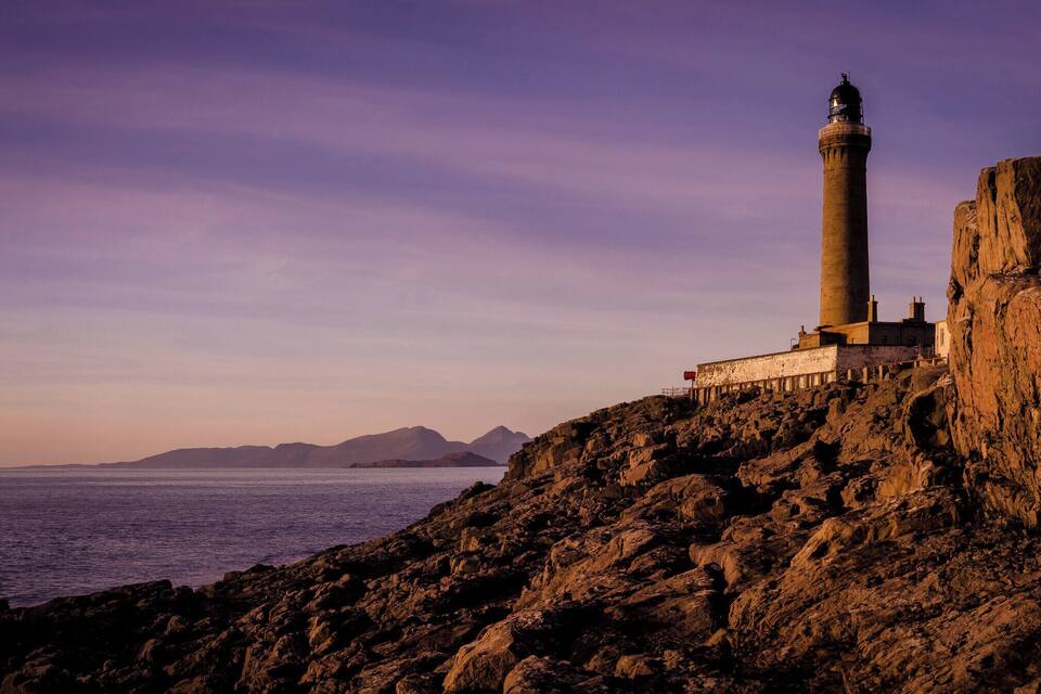 A lighthouse on a rocky coastline with a clear view of the sea and distant mountains.